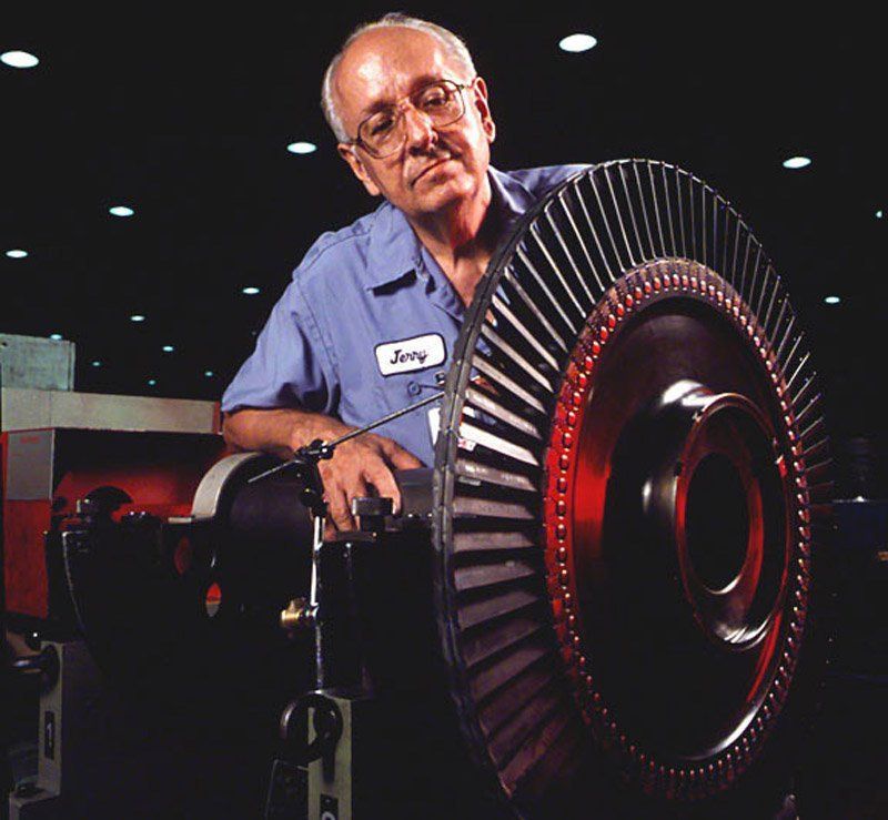 Technician in blue shirt inspects a large turbine engine in a dimly lit workshop.