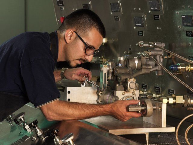 Man operating a machine in a workshop, leaning close to inspect a metal component
