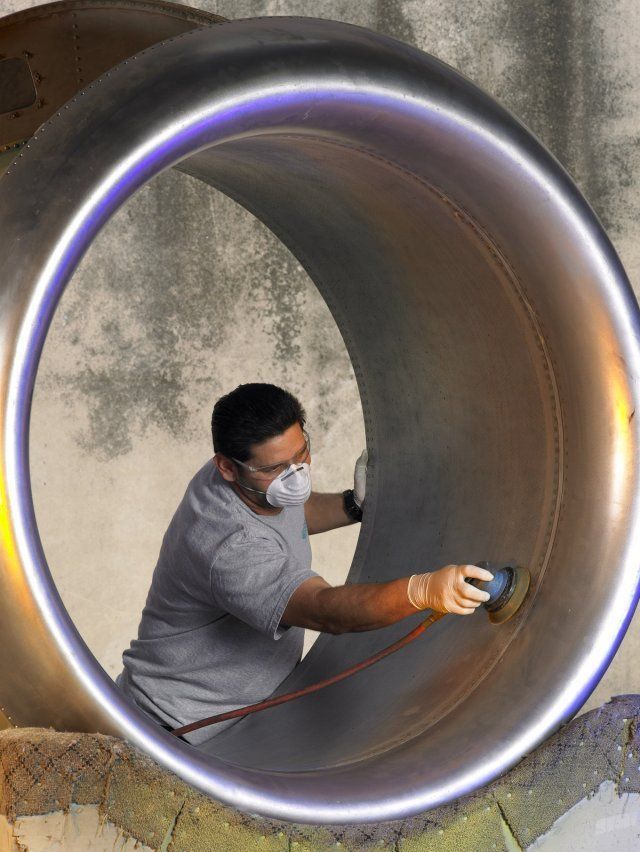 Worker in a mask and gloves cleaning a large metal pipe interior with a power tool.