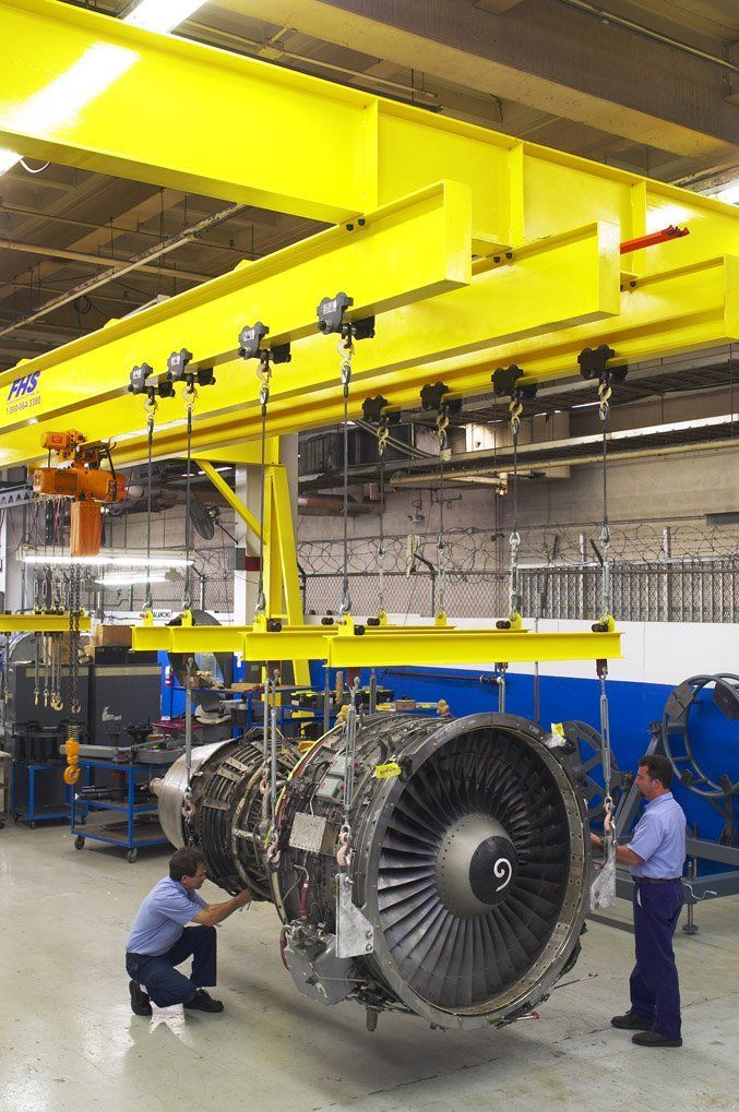 Workers inspecting a large jet engine suspended in a factory by a yellow overhead crane
