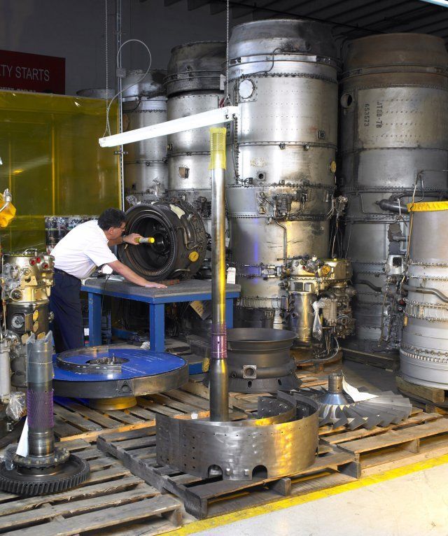 Worker in white shirt handling a large black wheel in an industrial storage area with stacked metal drums.
