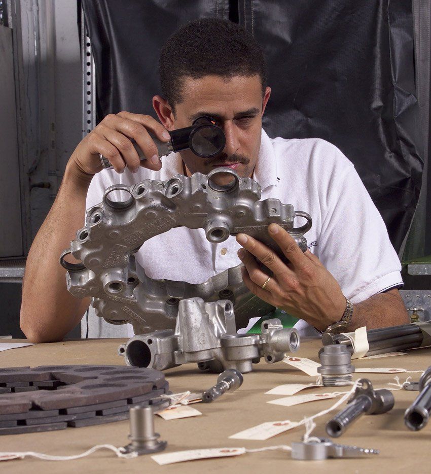 Technician inspecting a metal engine part with a magnifying glass at a workbench.