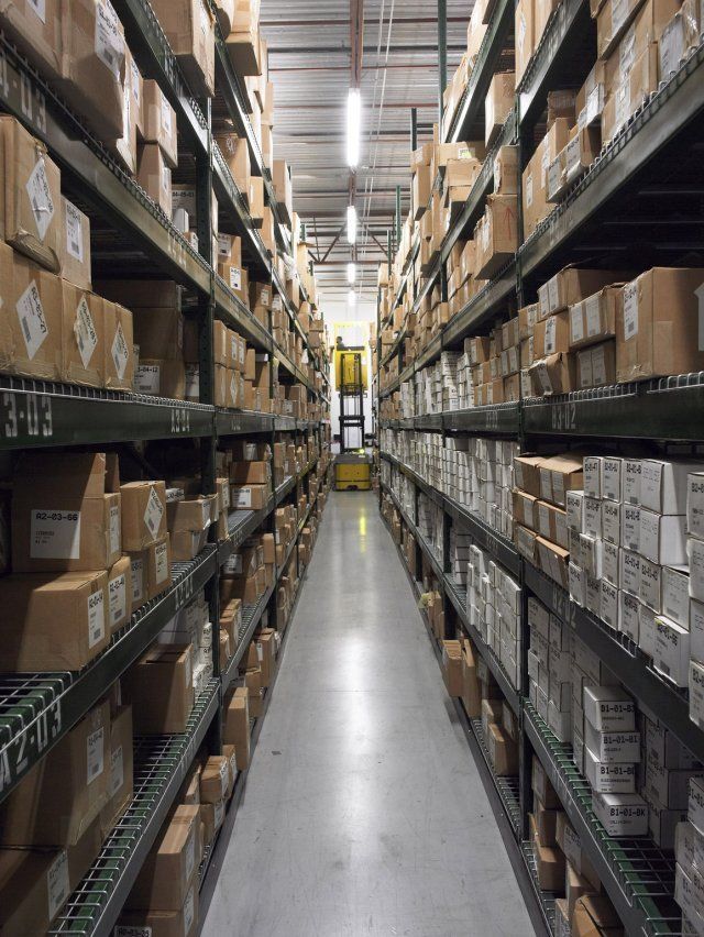 A narrow warehouse aisle lined with stacked boxes on tall shelves and bright overhead lights.