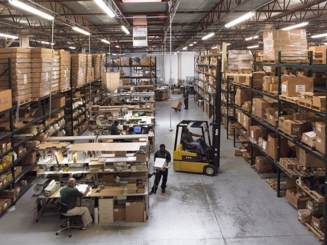 Busy warehouse with tall shelving, stacked boxes, and workers operating a forklift and packing tables.