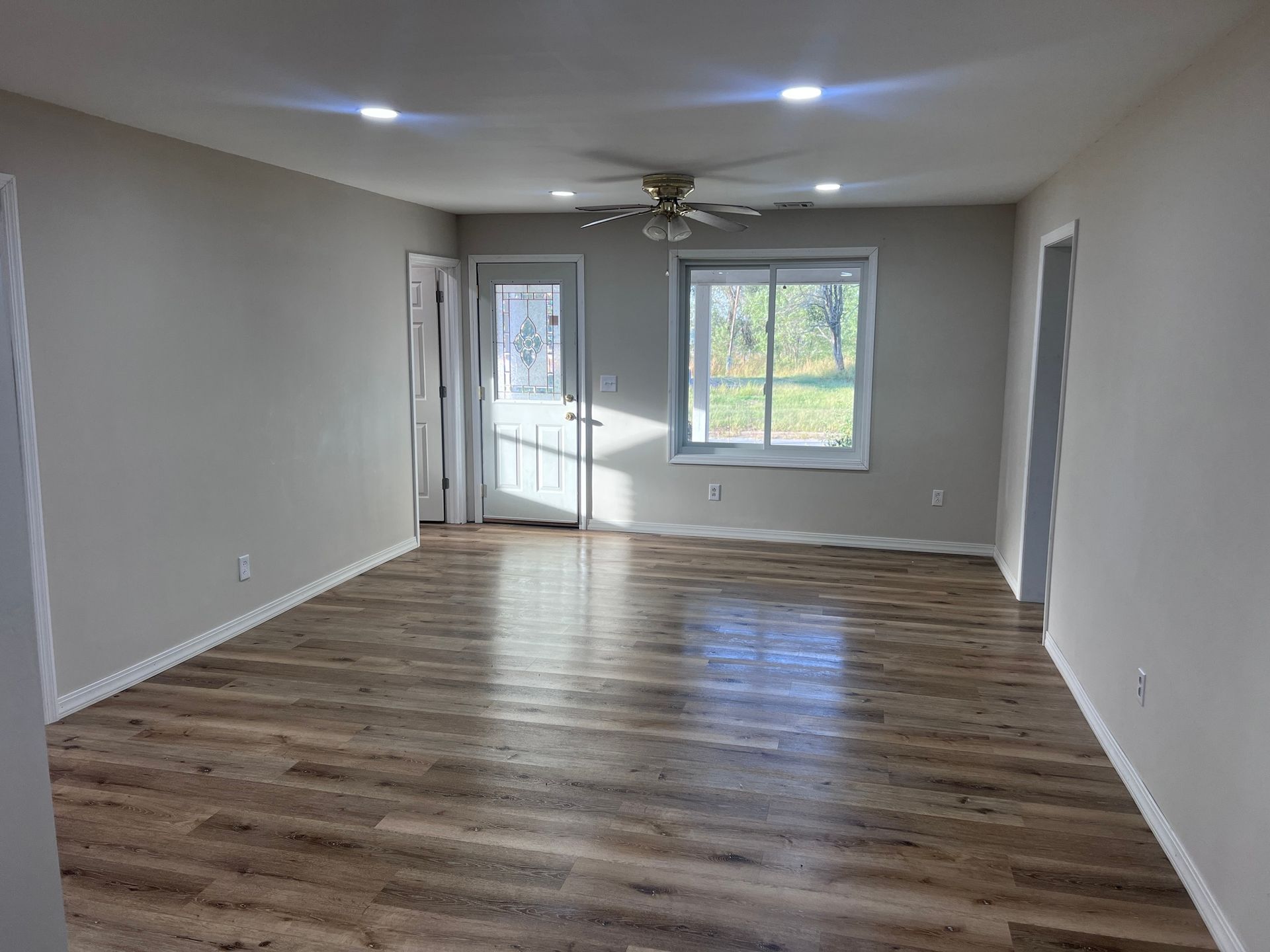 Empty living room with wood-look flooring, door, and window. Light gray walls, ceiling fan, and recessed lights.