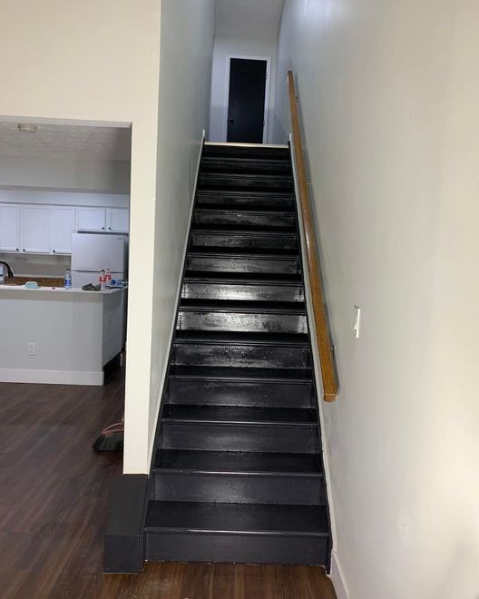 Black painted staircase leading upwards with a wooden handrail on the right. White walls, kitchen in background.