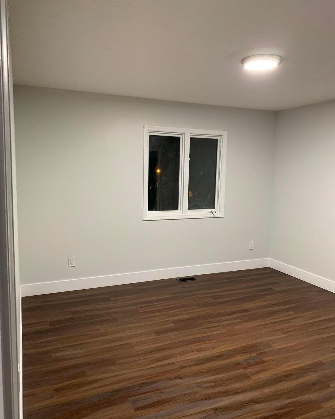 Empty room with gray walls, white trim, and dark wood-look flooring. A window and ceiling light are visible.