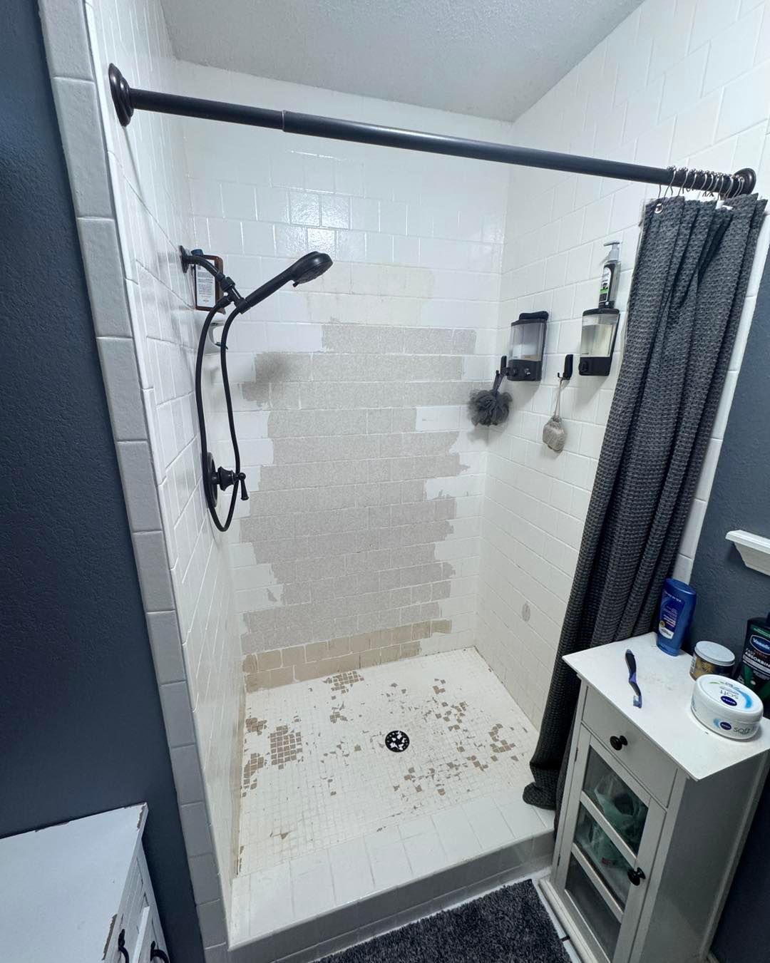 Shower stall with exposed tile, showerhead, curtain rod, and small white cabinet.
