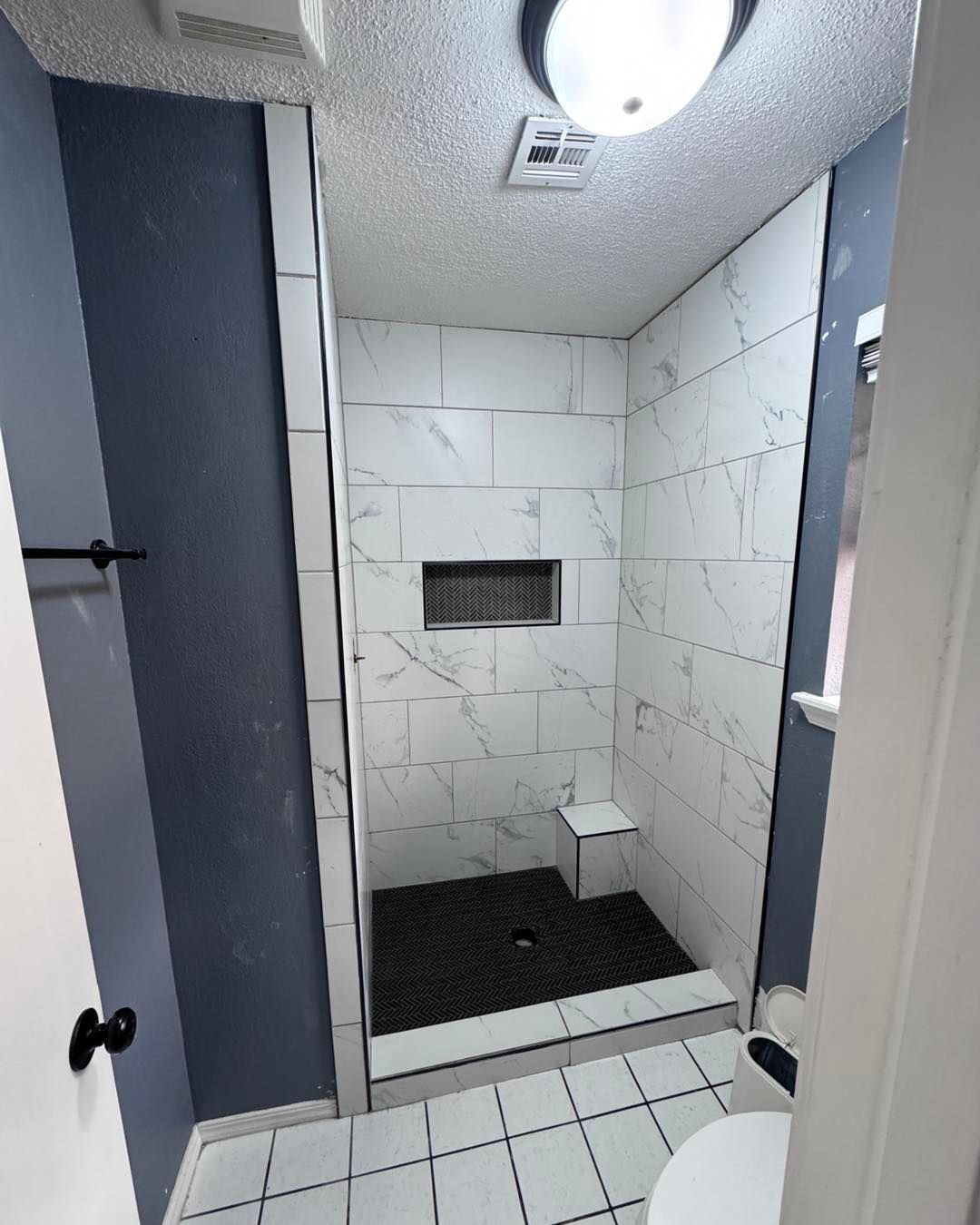 A newly remodeled shower with white marble-look tiles, a black floor, a built-in bench, and a dark blue wall.