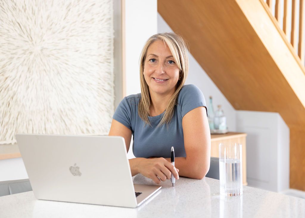 Woman smiling at a white laptop on a bright desk with a glass of water and wooden staircase behind her