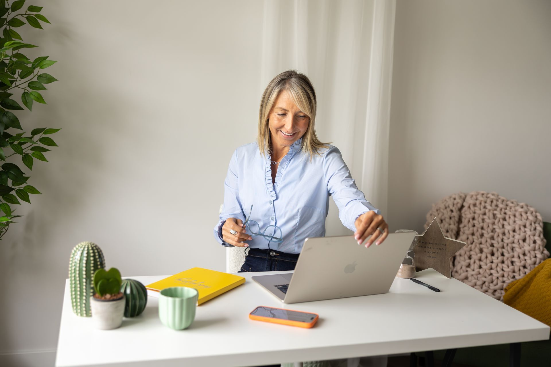 Woman at a white desk using a laptop, with notebooks, a mug, and small plants nearby.