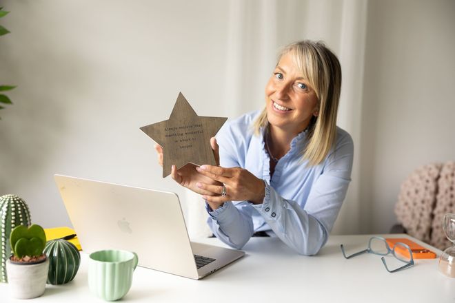 Woman smiling at desk, holding a star-shaped plaque beside a laptop and coffee mugs
