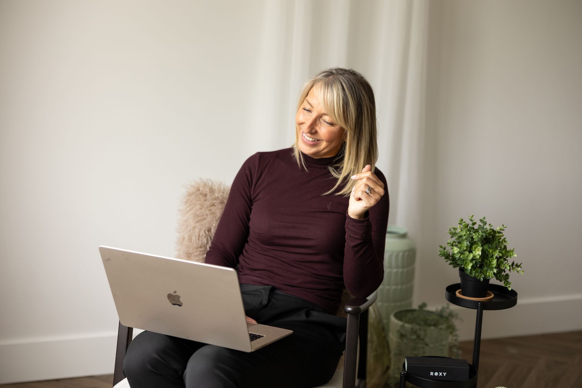 Woman smiling with a laptop on her lap in a bright room, seated beside a small plant on a side table
