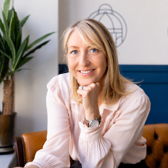 Smiling blonde woman in a pale pink blouse resting her chin on her hand in an office setting