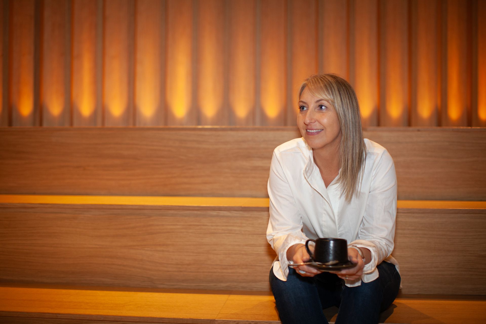 Woman seated on a wooden bench holding a black cup in a warmly lit room