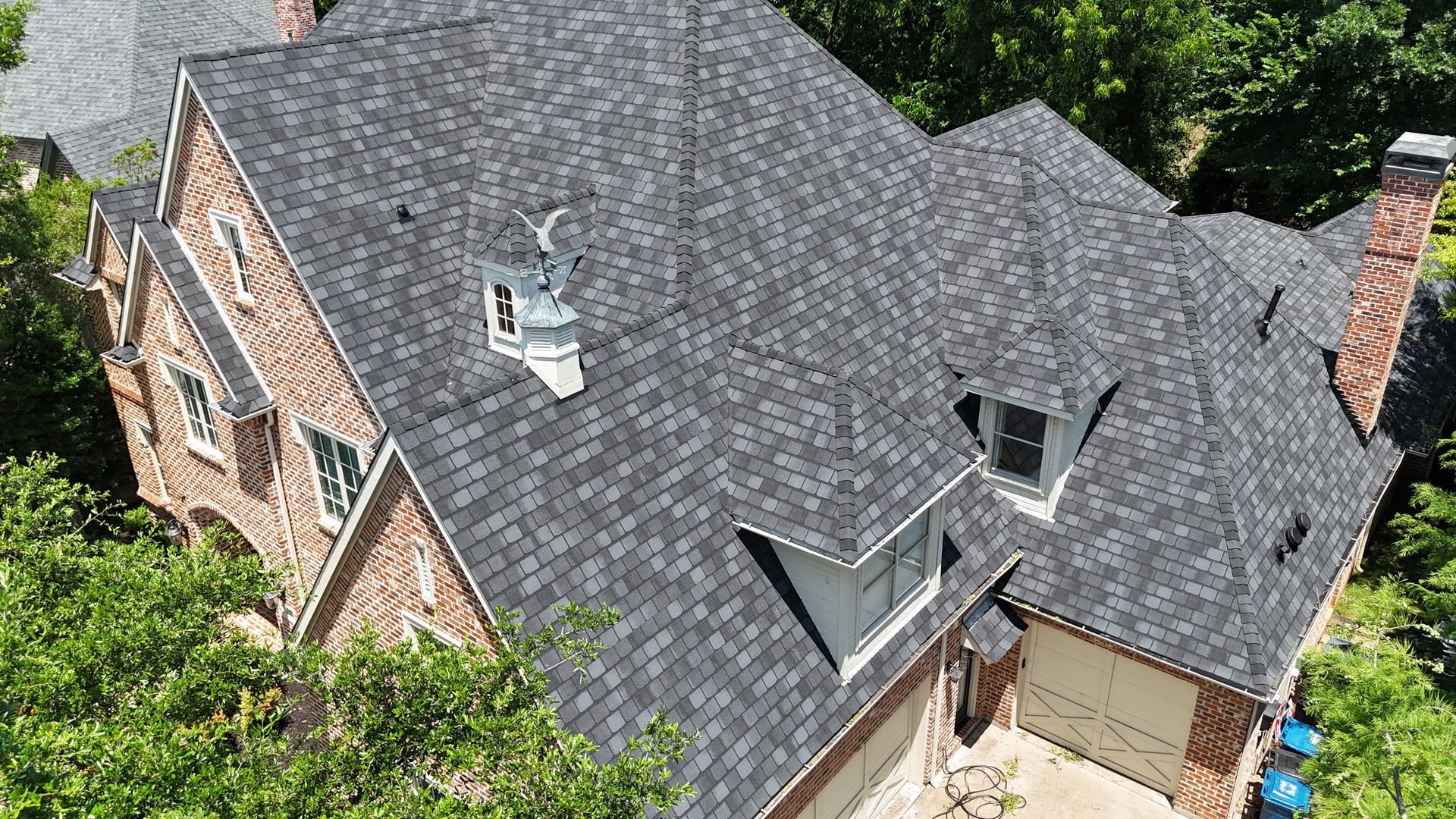 Gray asphalt shingle roof on a two-story brick house with dormer windows, surrounded by trees.