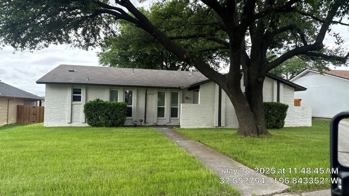 White house with gray roof, green lawn, and large tree in front.