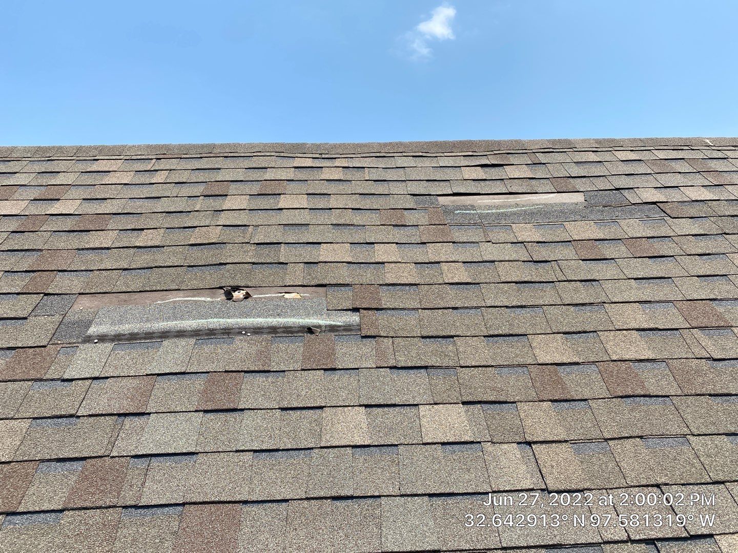 Close-up of a damaged shingle roof under a blue sky, showing missing and broken shingles.