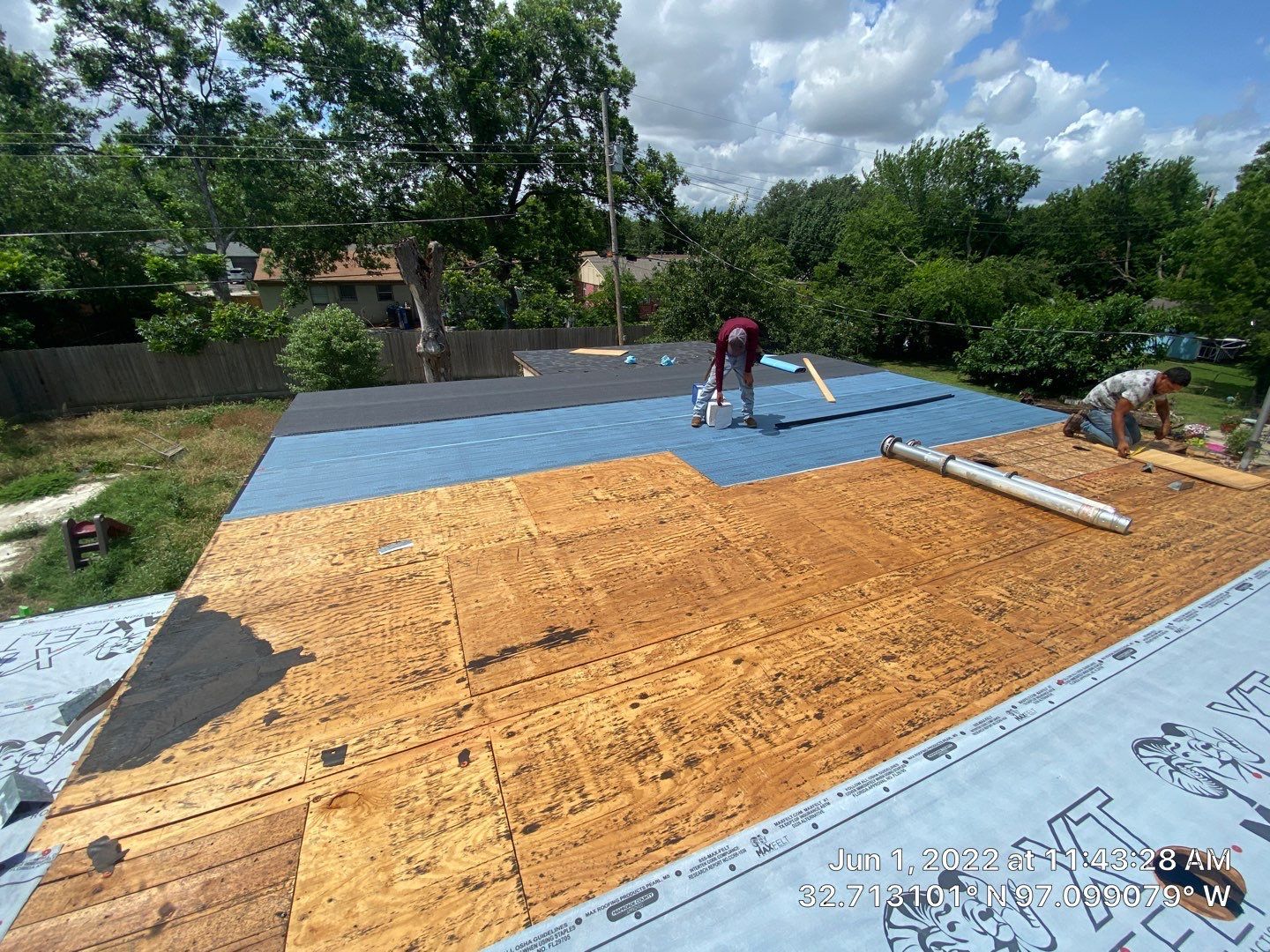 Roofers installing roofing material on a blue-covered roof, sunny day, surrounded by trees.