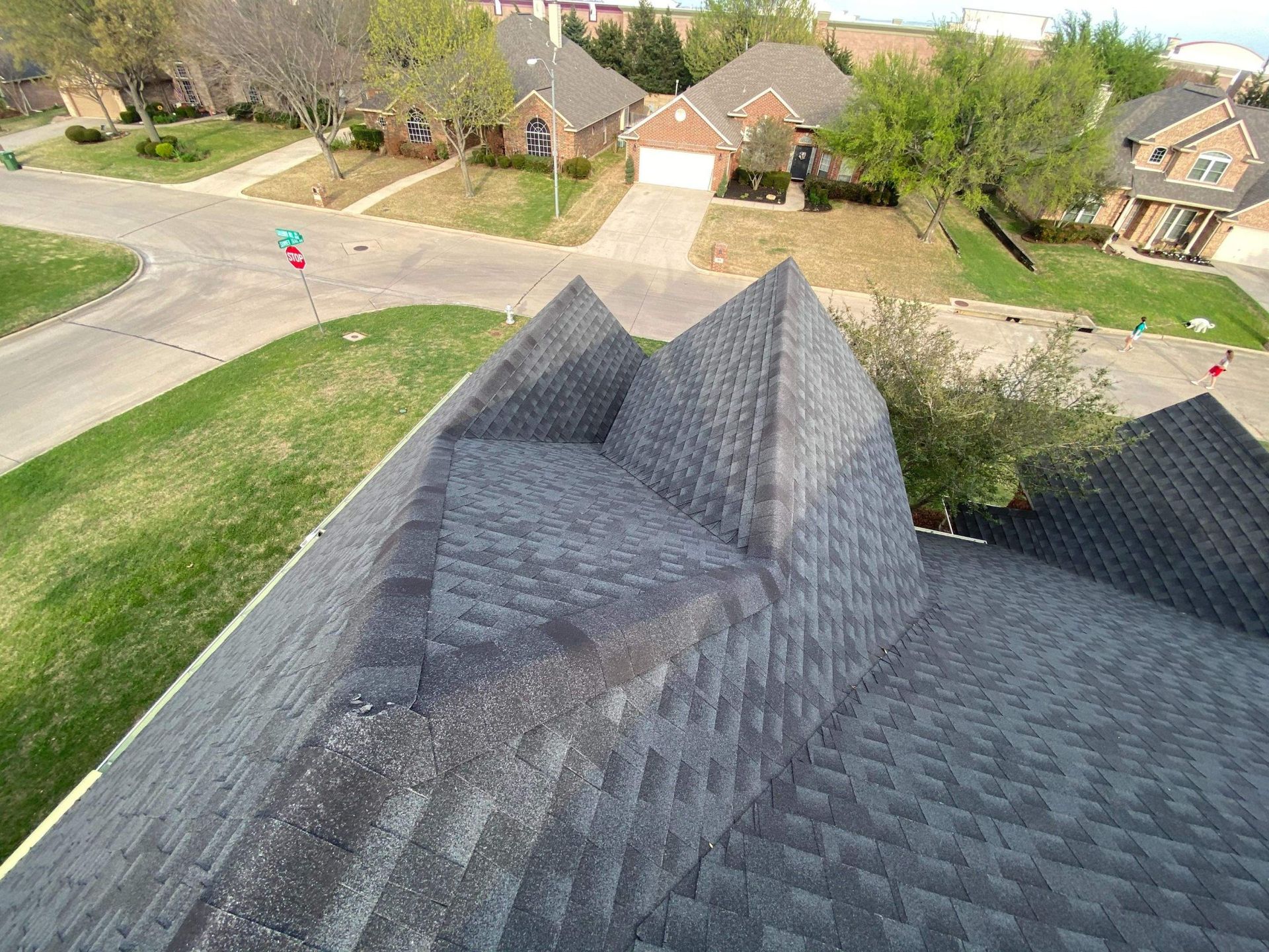 Overhead view of a house roof with shingles. Houses and a street are visible in the background. Green grass surrounds.