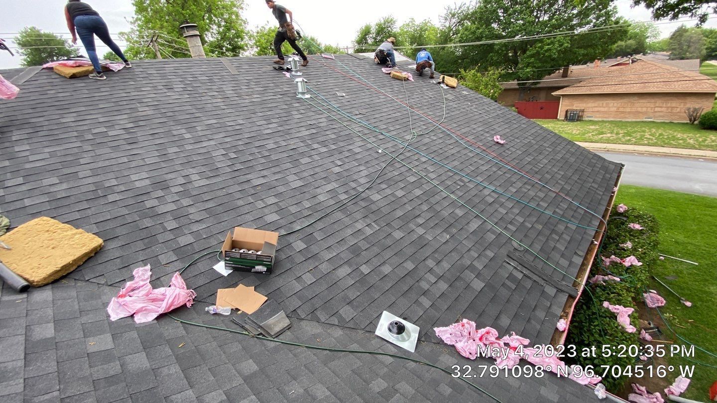 Roofers working on a residential roof, shingles being laid. Green lawn, trees, blue sky.