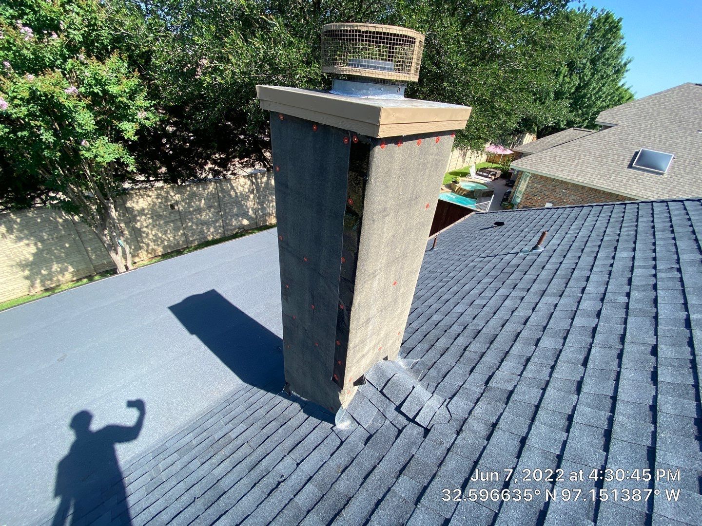 Chimney on a roof. A person's shadow is visible. Blue sky and foliage in the background.