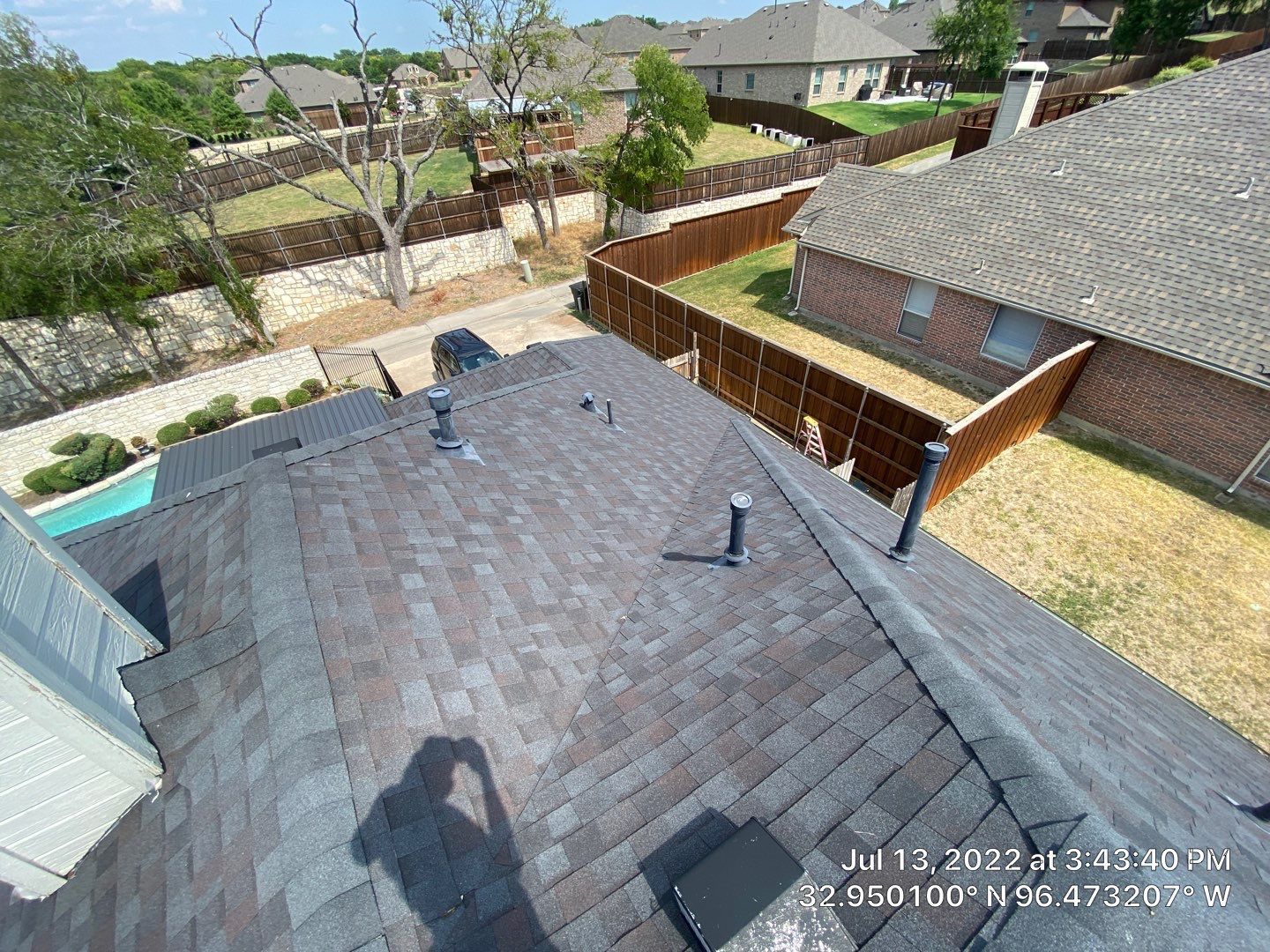 Aerial view of a dark asphalt shingle roof. Other houses and yards are visible beyond the roof.