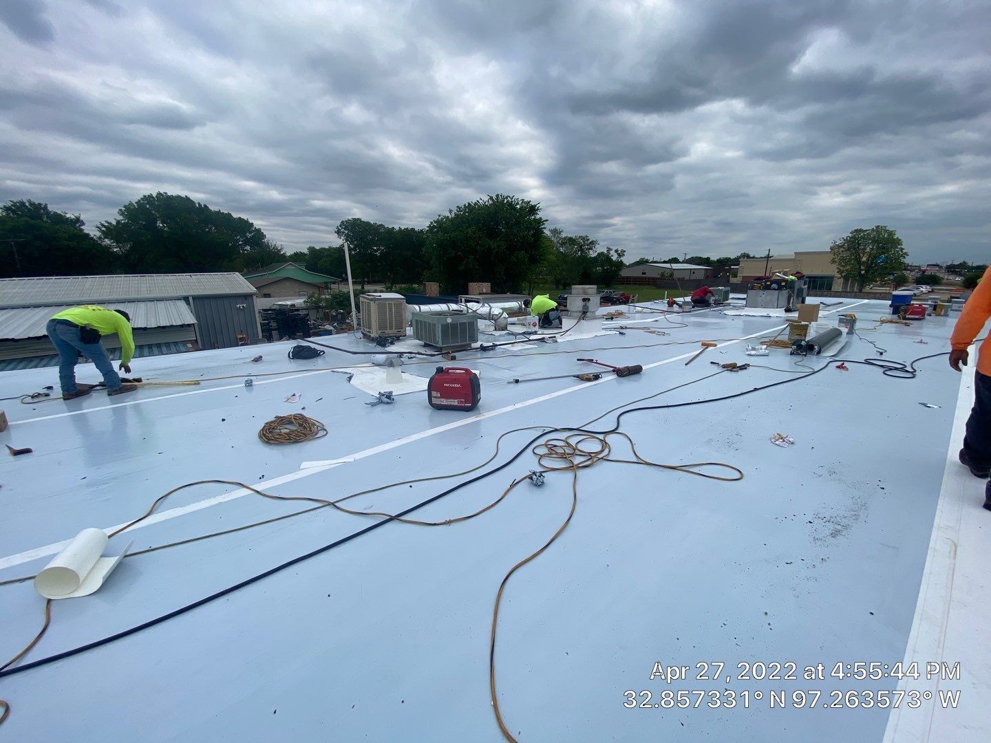 Construction workers on a blue flat roof, working near equipment and power cords, cloudy sky.