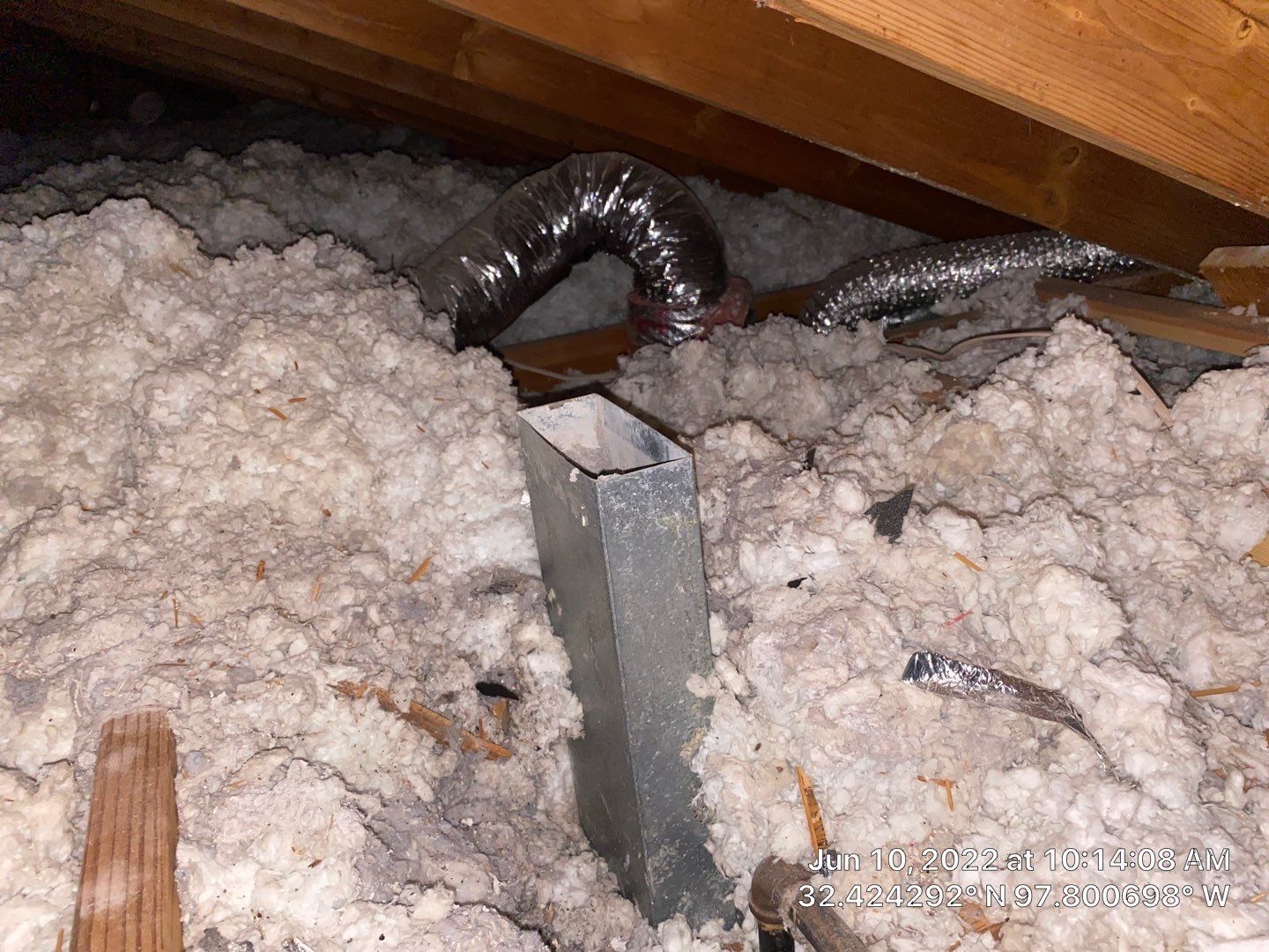 Attic with insulation, a metal duct, and flexible silver ductwork. Brown wooden rafters are visible.
