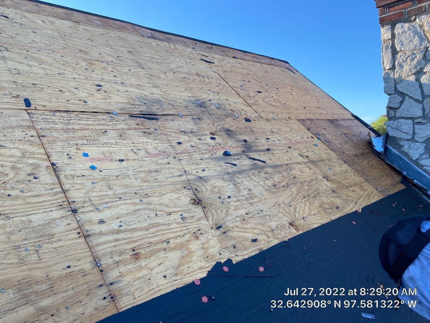Exposed roof with plywood sheathing, dark asphalt shingles, and stone chimney against a blue sky.