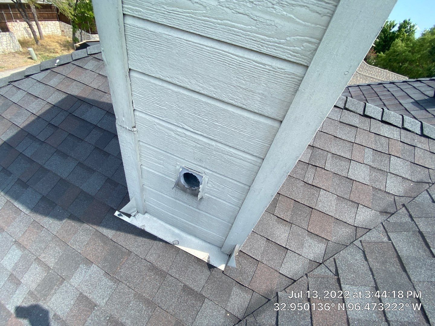 Chimney on a roof. Brown shingles surround a white and grey structure with a small hole.