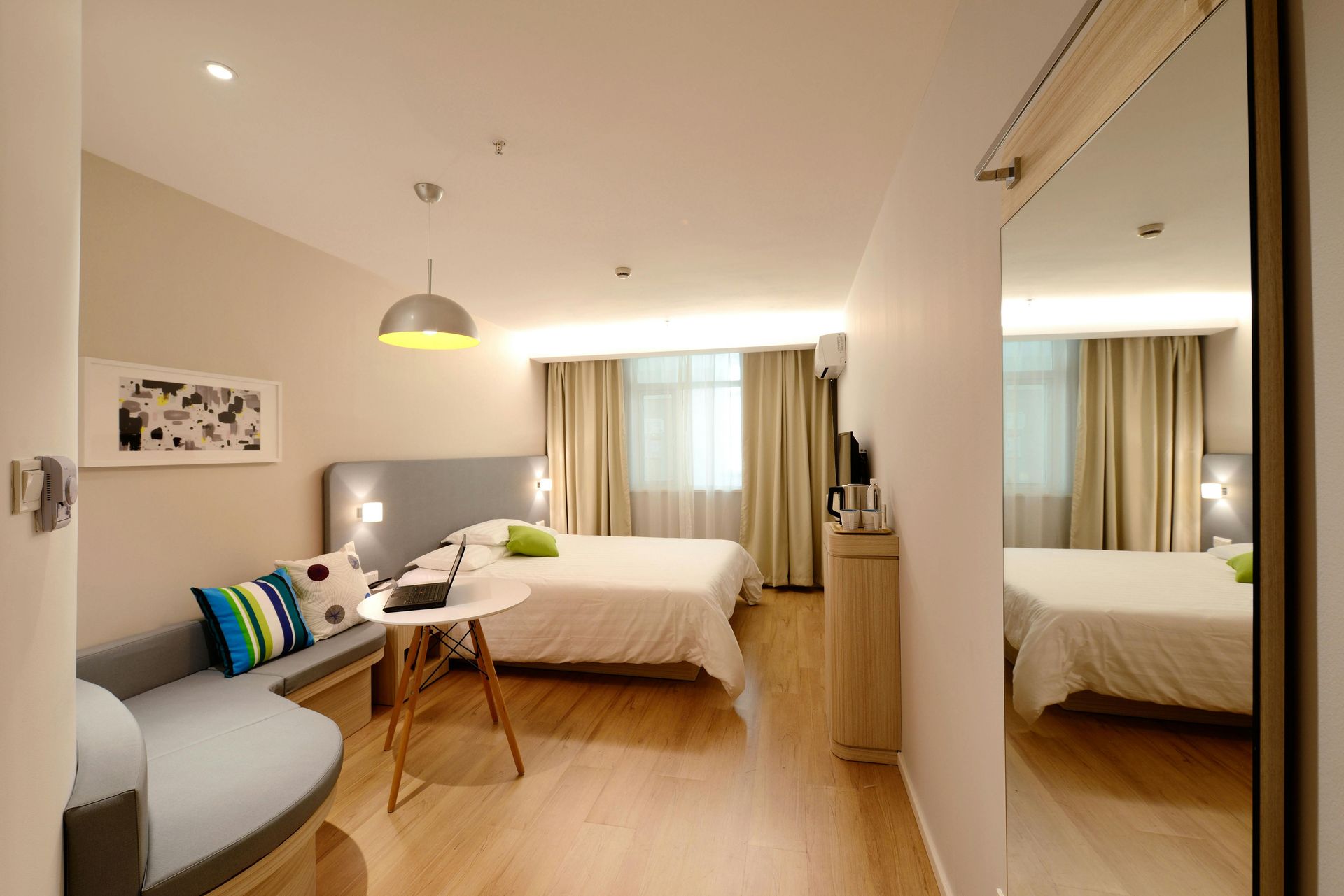 Hotel room with a bed, small sofa, and a table. A large mirror reflects the room.