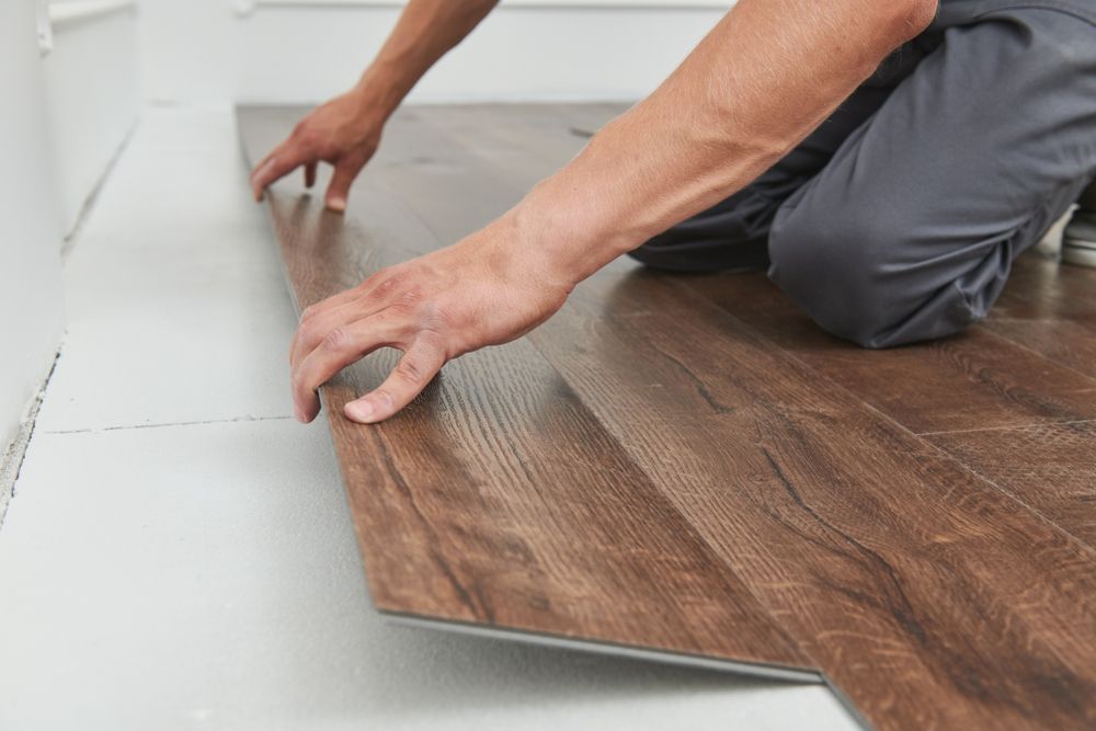 Person installing wood-look flooring on a white subfloor, indoors.