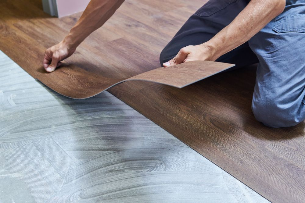 Person installing brown wood-look flooring over a light gray floor, indoors.