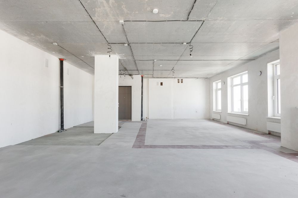 Empty, unfinished interior space with white walls, concrete ceiling and floor, and windows.