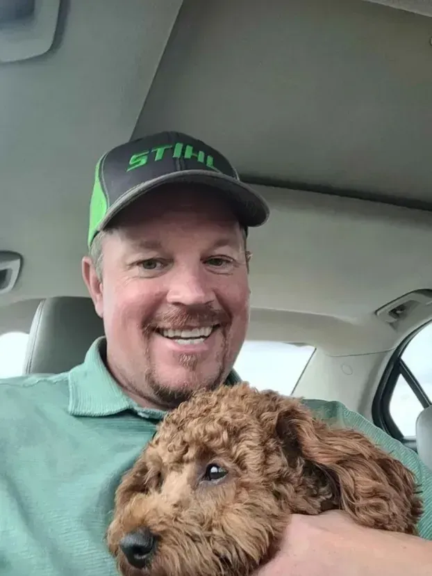 Man in a Stihl hat smiling, holding a brown Goldendoodle puppy inside a car.