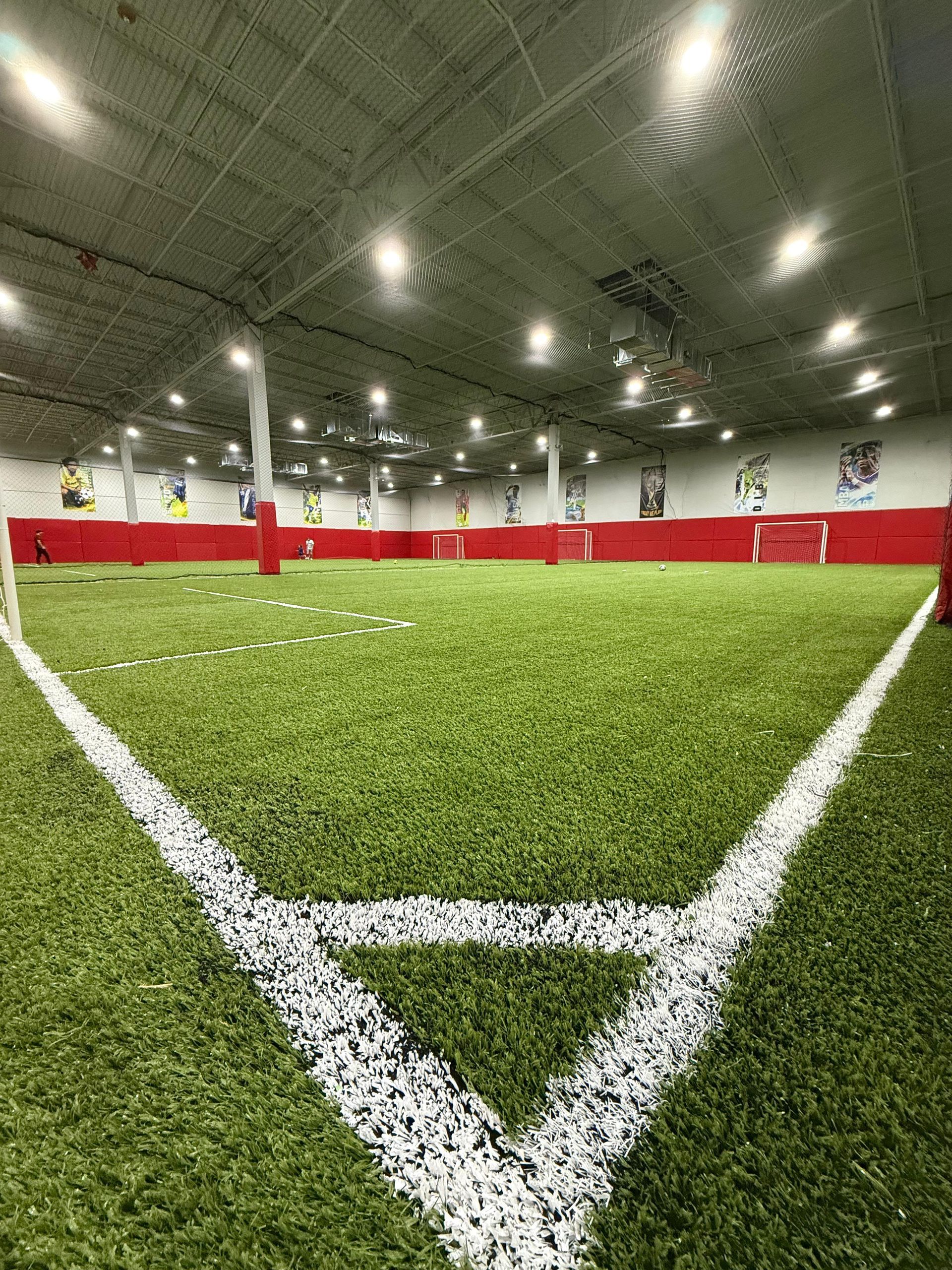 Indoor turf field with white corner markings, red barriers, and overhead lighting.