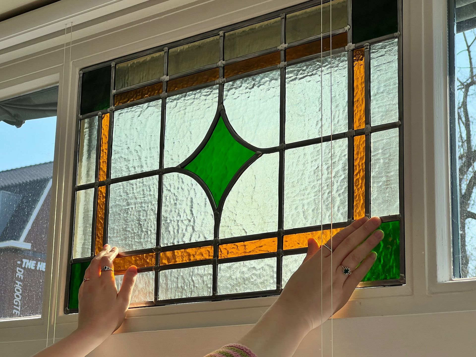 Twee handen houden een glas-in-loodpaneel met een groene ruit tegen het raam