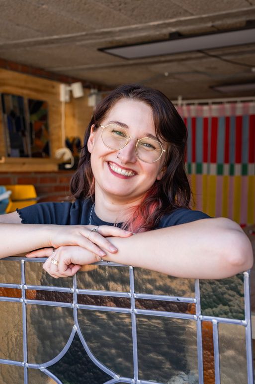 Lieke Kleverwal lacht, leunend op een glas-in-loodraam in haar atelier