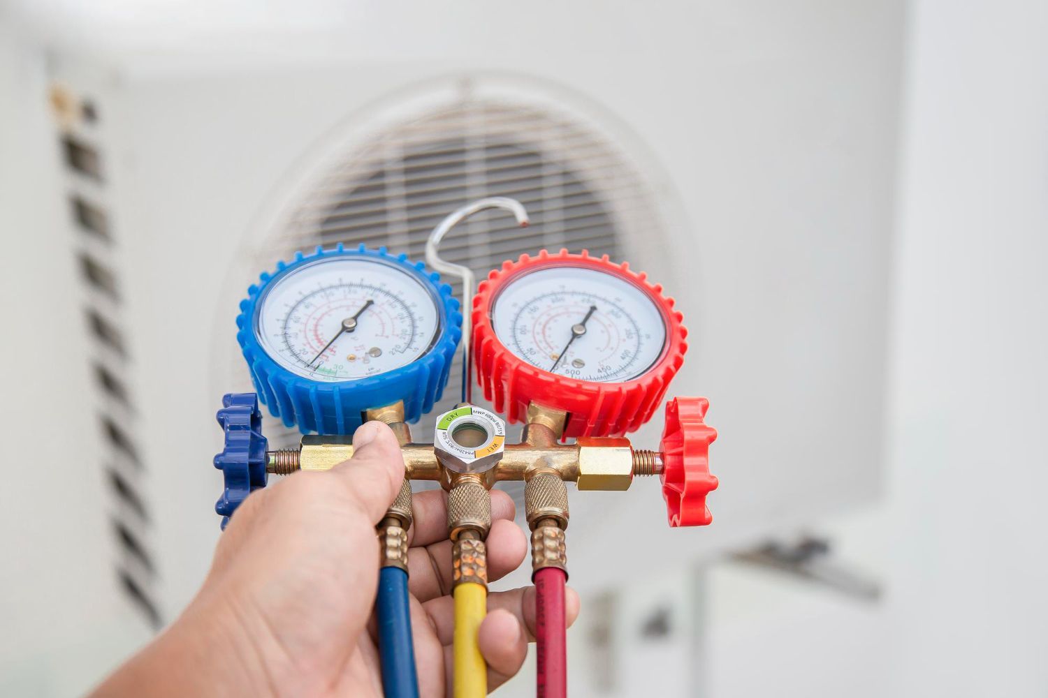 Hand holding blue and red HVAC pressure gauges in front of a fan vent