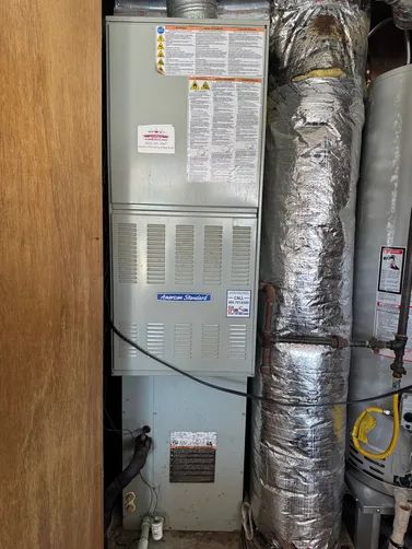 Furnace and ductwork in a utility closet, with silver insulated pipes and a gray metal panel.