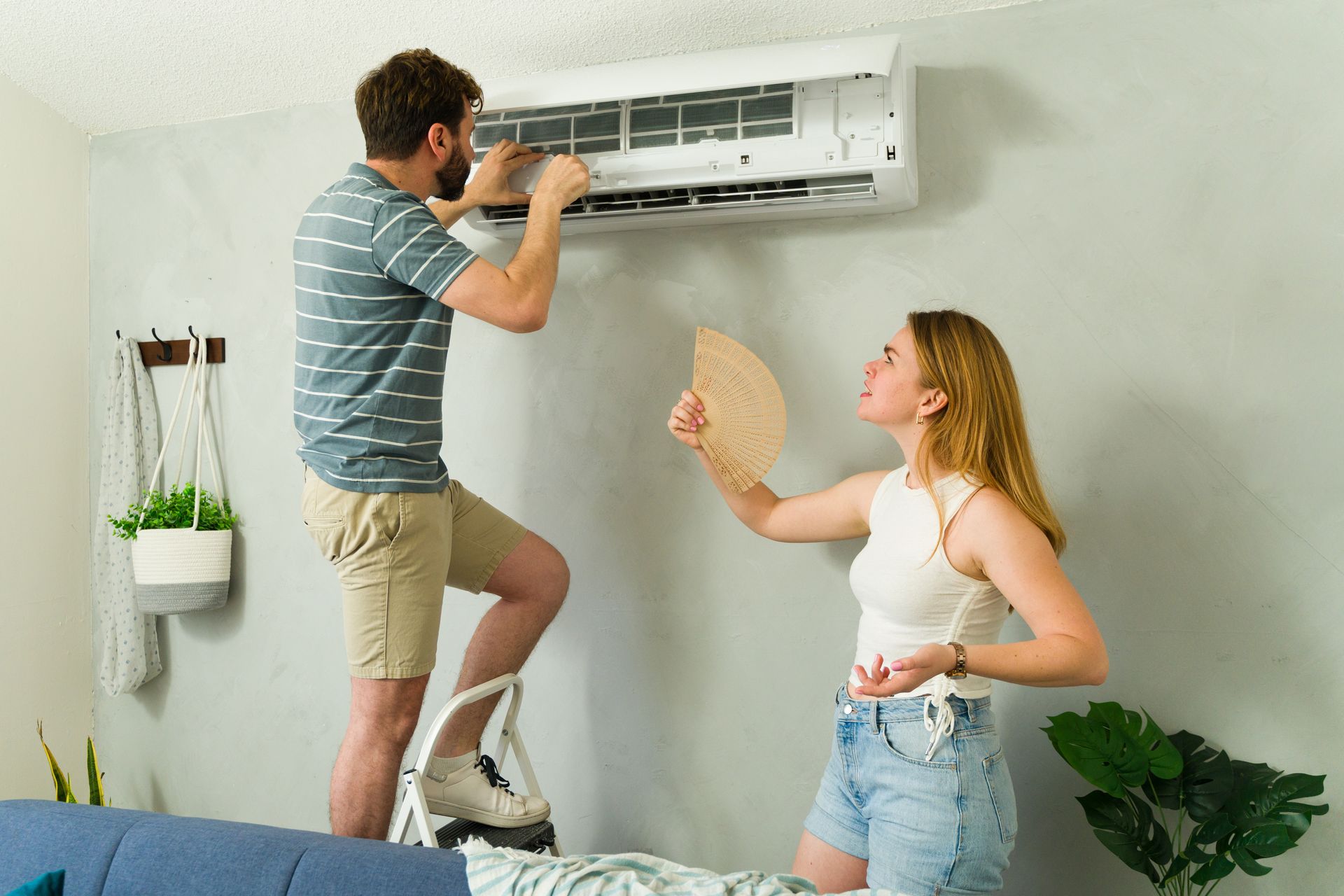 A person on a step stool repairs a wall-mounted air conditioner while another person holds a fan nearby.
