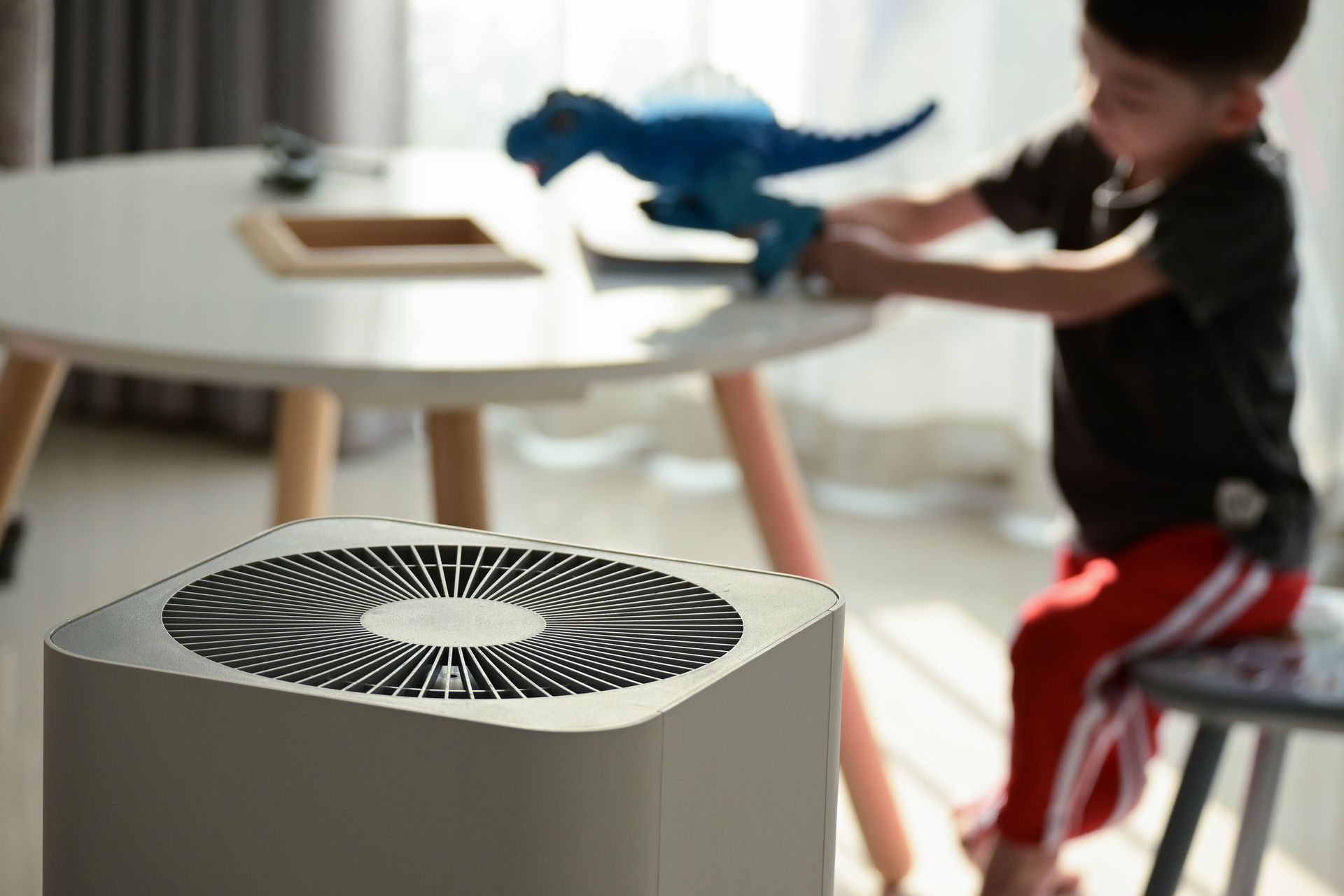 A white air purifier stands in the foreground as a person plays with a blue toy dinosaur at a small table nearby.