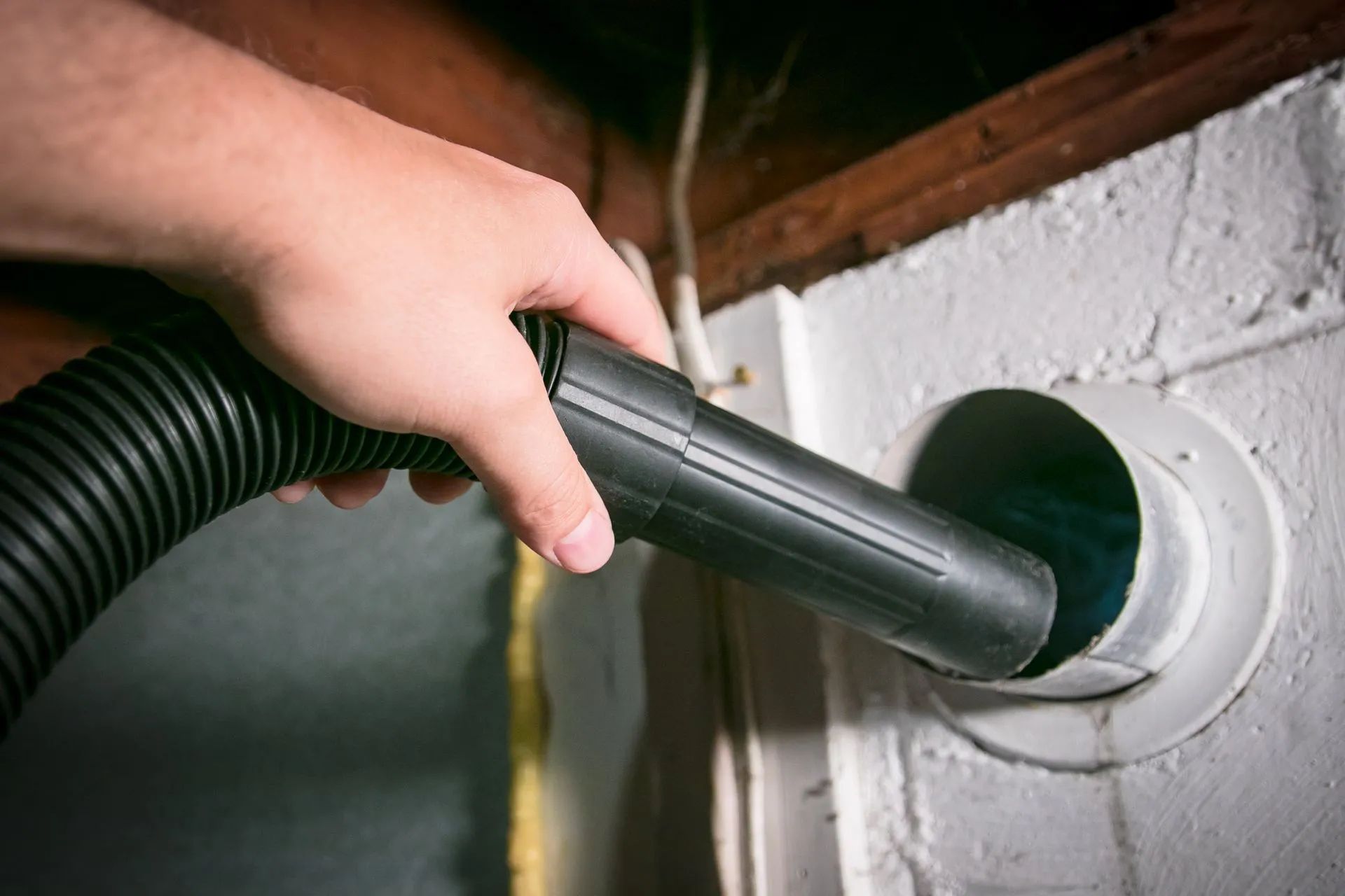 Hand holding a vacuum hose inserted into a white wall vent opening