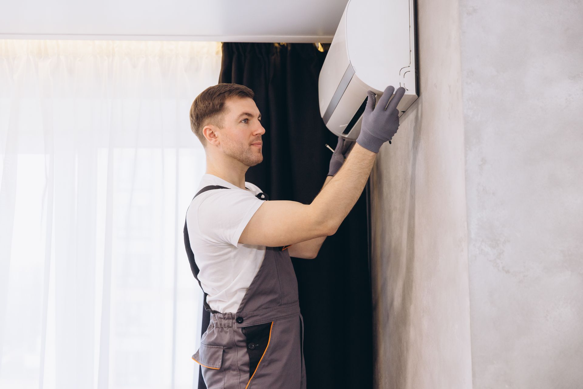 A technician wearing grey work coveralls and gloves mounts or repairs a white wall-mounted air conditioning unit.