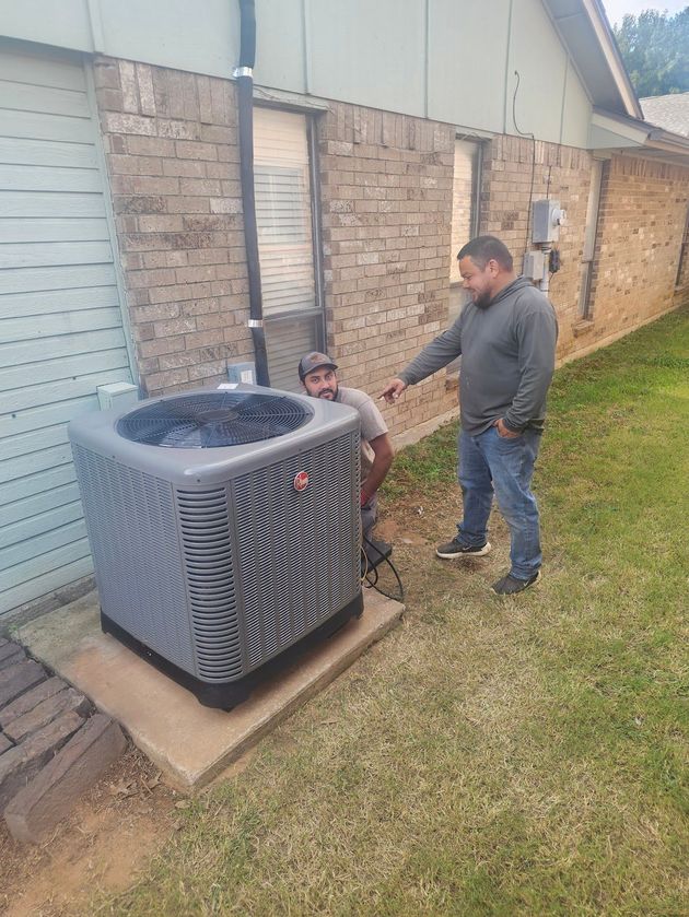 Two men inspecting an air conditioning unit outside a brick building. One points at it.