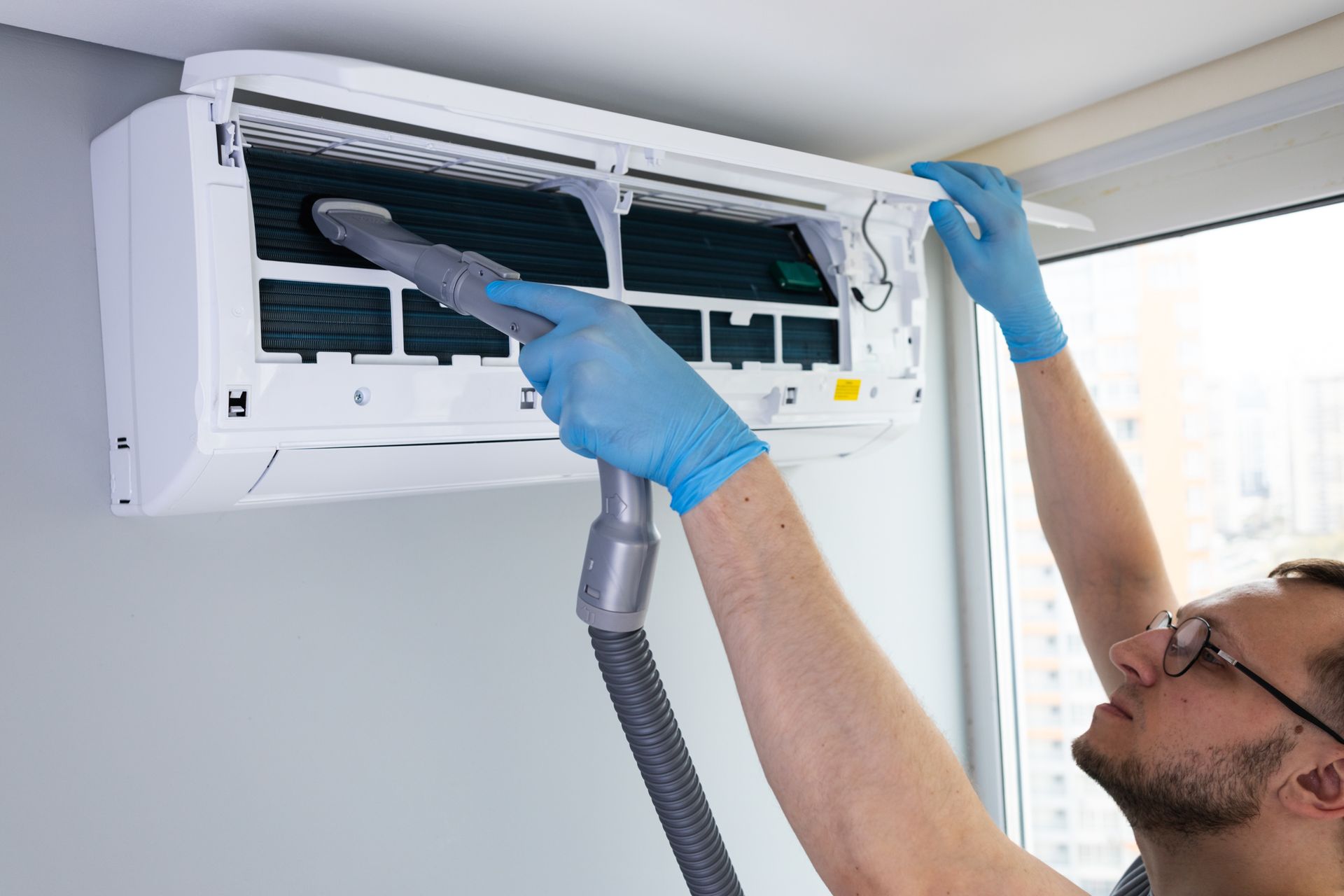 A technician wearing blue gloves uses a vacuum attachment to clean the dust filter of a wall-mounted air conditioner.