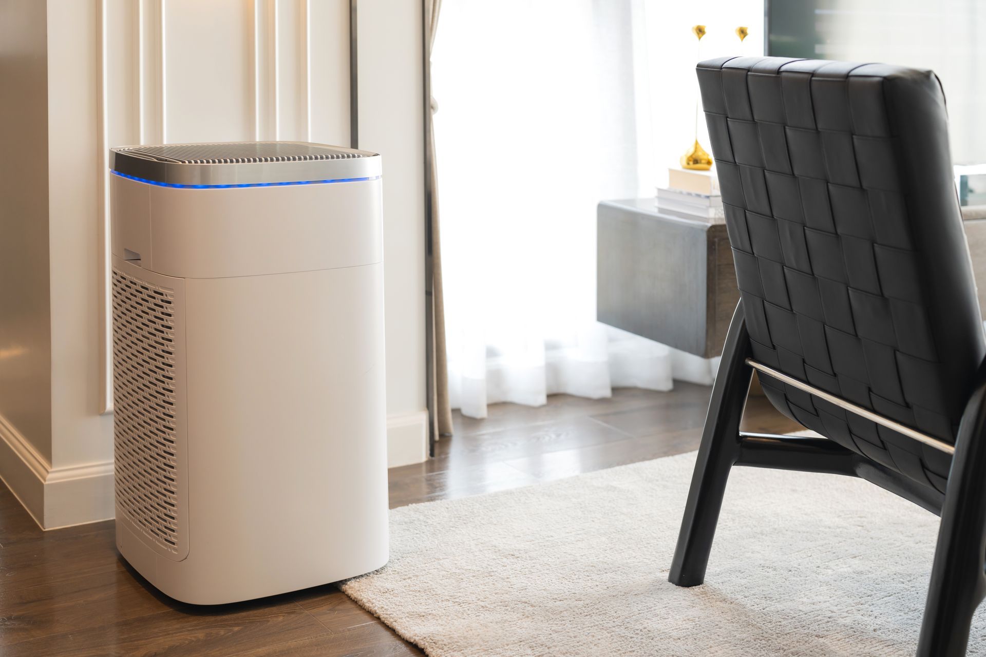 A white air purifier with a glowing blue light stands on a wooden floor next to a woven black chair in a modern room.