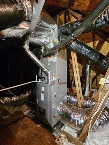 Attic HVAC furnace with silver ductwork and insulation in a dim, unfinished crawlspace