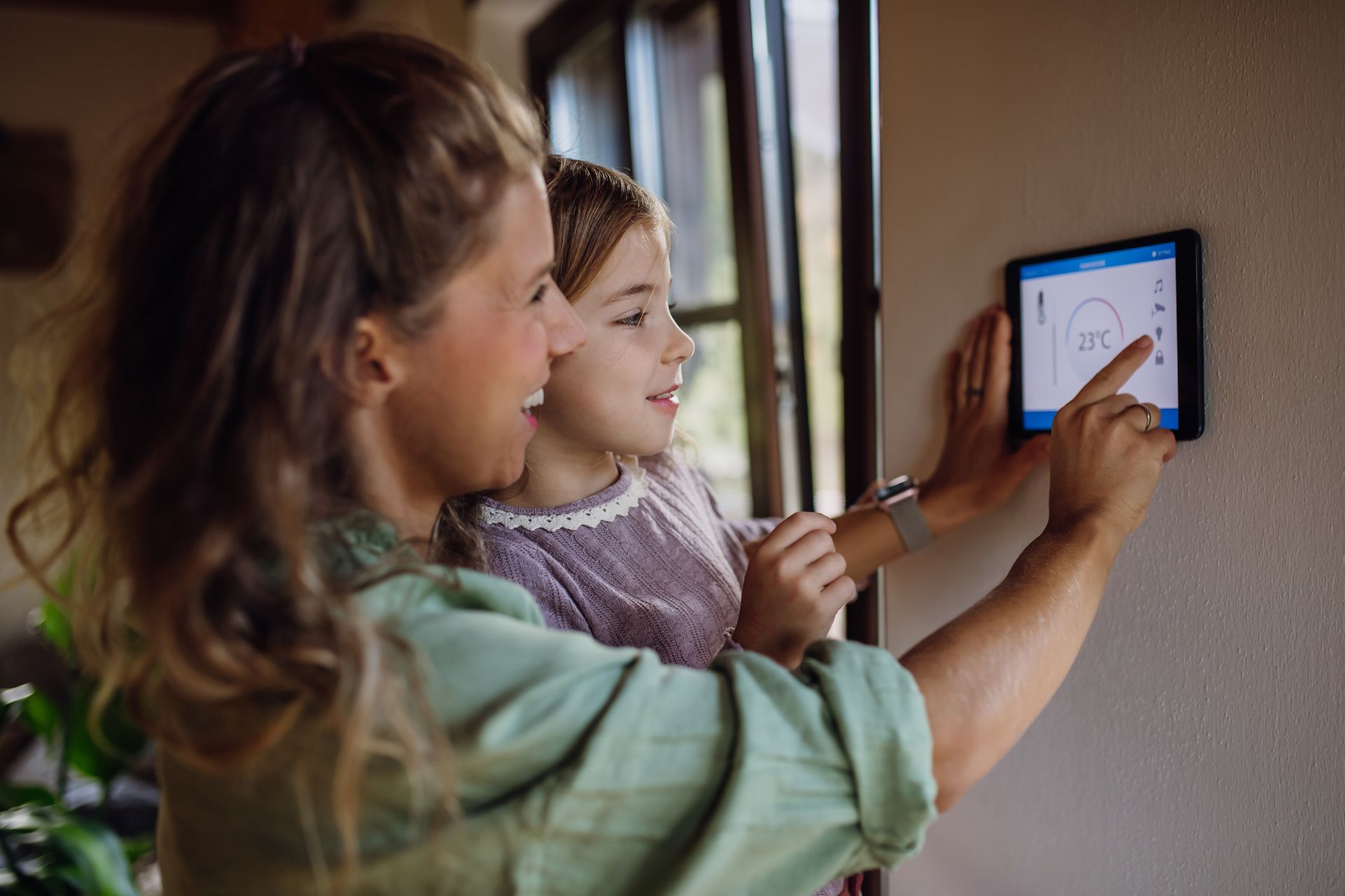 A person and a child touch a smart home wall tablet to adjust room temperature settings in a home.
